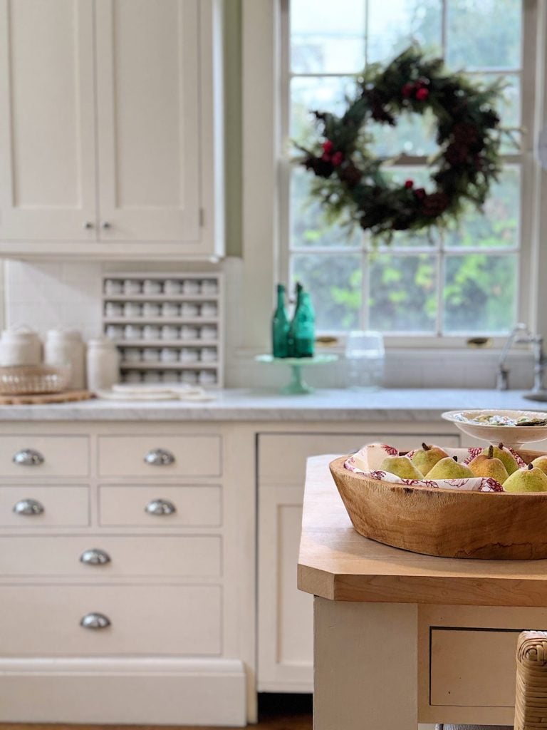 A kitchen scene with a wooden bowl of pears on an island, white cabinetry, and a wreath hanging in the window above the sink.