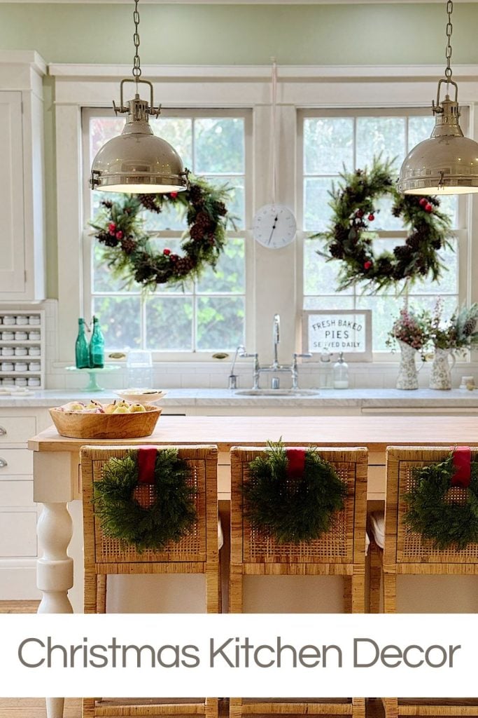 A kitchen with two hanging lights, wreaths on the window and chairs, wooden stools, and a counter with a bowl. Text on image says "Christmas Kitchen Decor.