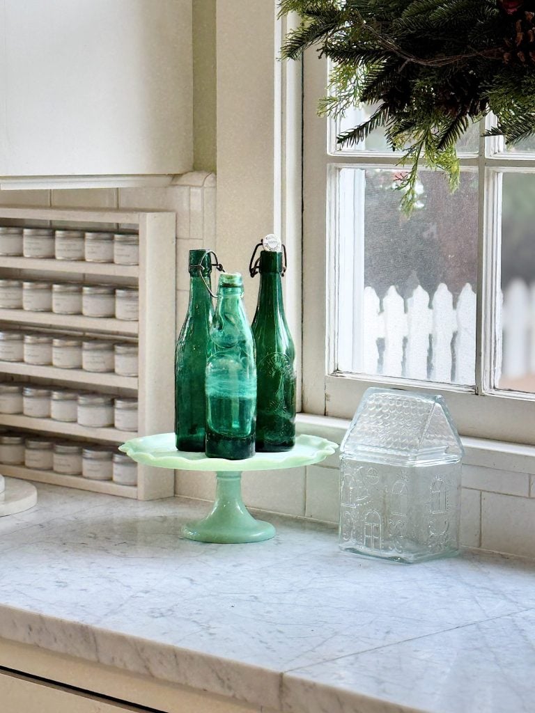 Green glass bottles on a jade pedestal stand next to a glass house decoration on a marble countertop near a window with greenery outside.