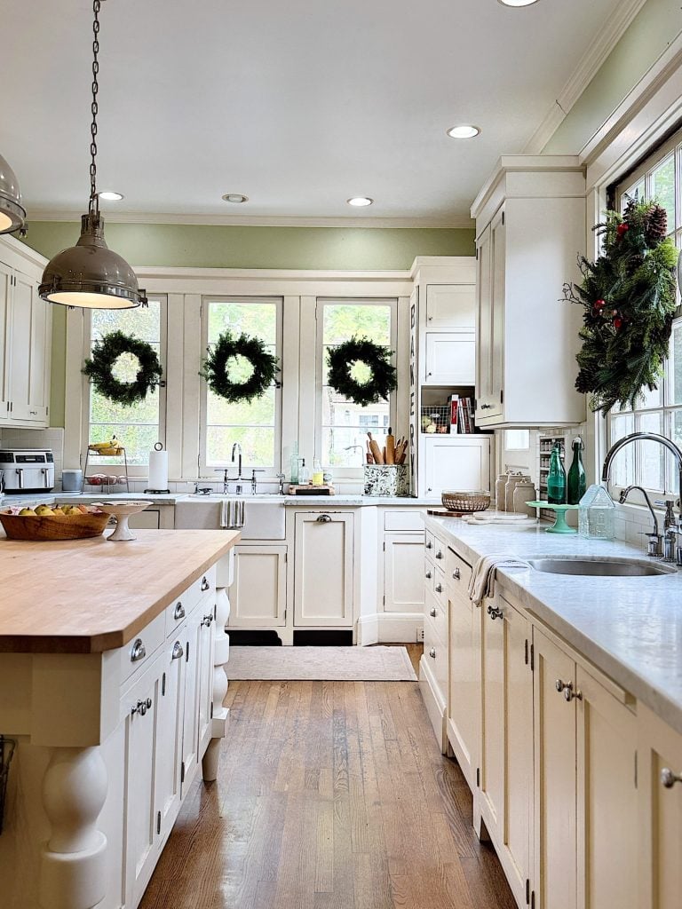 Bright kitchen with white cabinets, wooden floors, and a central island. Three wreaths hang on windows, and festive decorations accent the room.