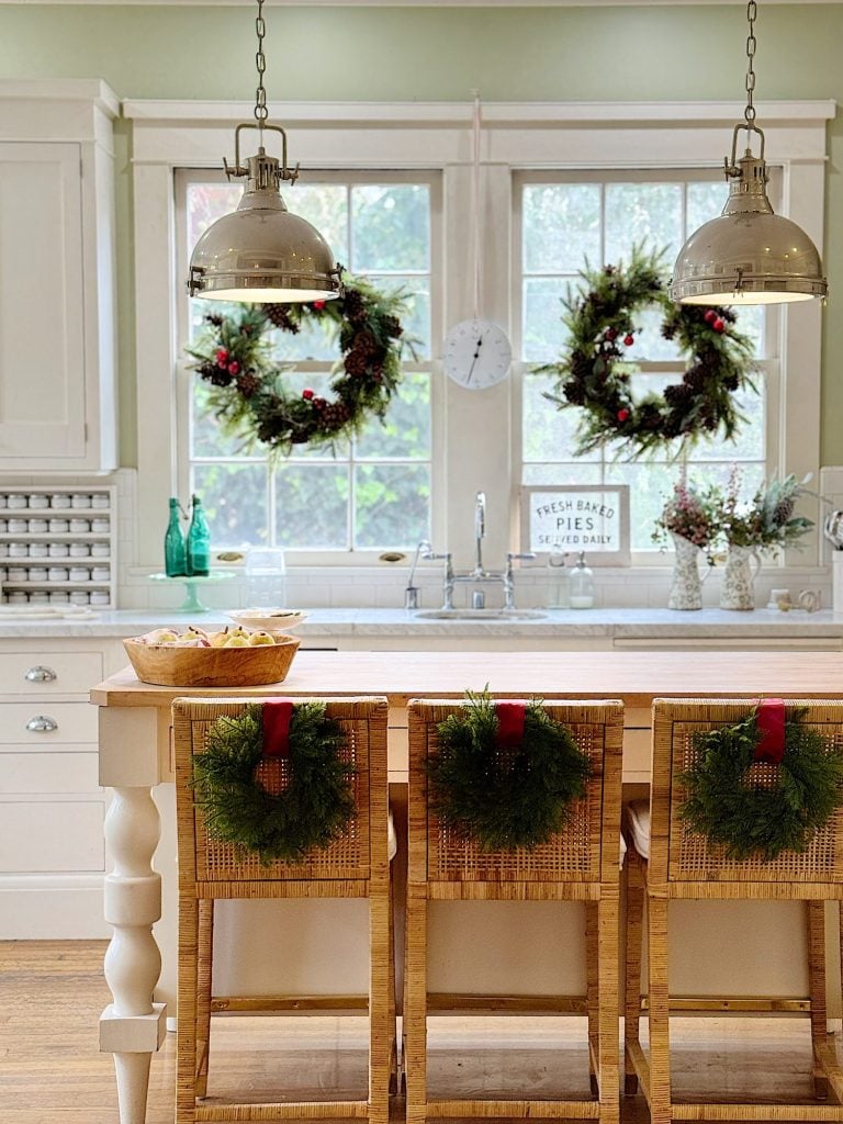 Kitchen with a wooden island, three wicker chairs adorned with wreaths, overhead pendant lights, and windows decorated with wreaths. A "Fresh Baked Pies" sign is visible on the counter.