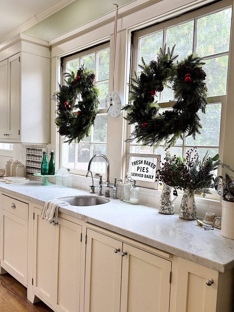 A kitchen with a white marble countertop, two wreaths on windows, and a sign reading "Fresh Baked Pies Served Daily" next to a sink.