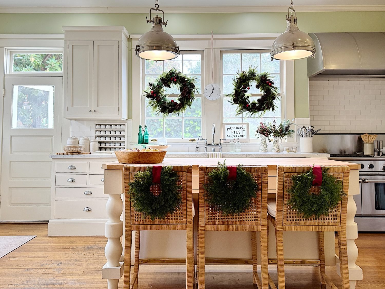 Bright kitchen with Christmas wreaths on windows and chairs, white cabinetry, and stainless steel appliances. Inviting holiday decor enhances the cozy atmosphere.