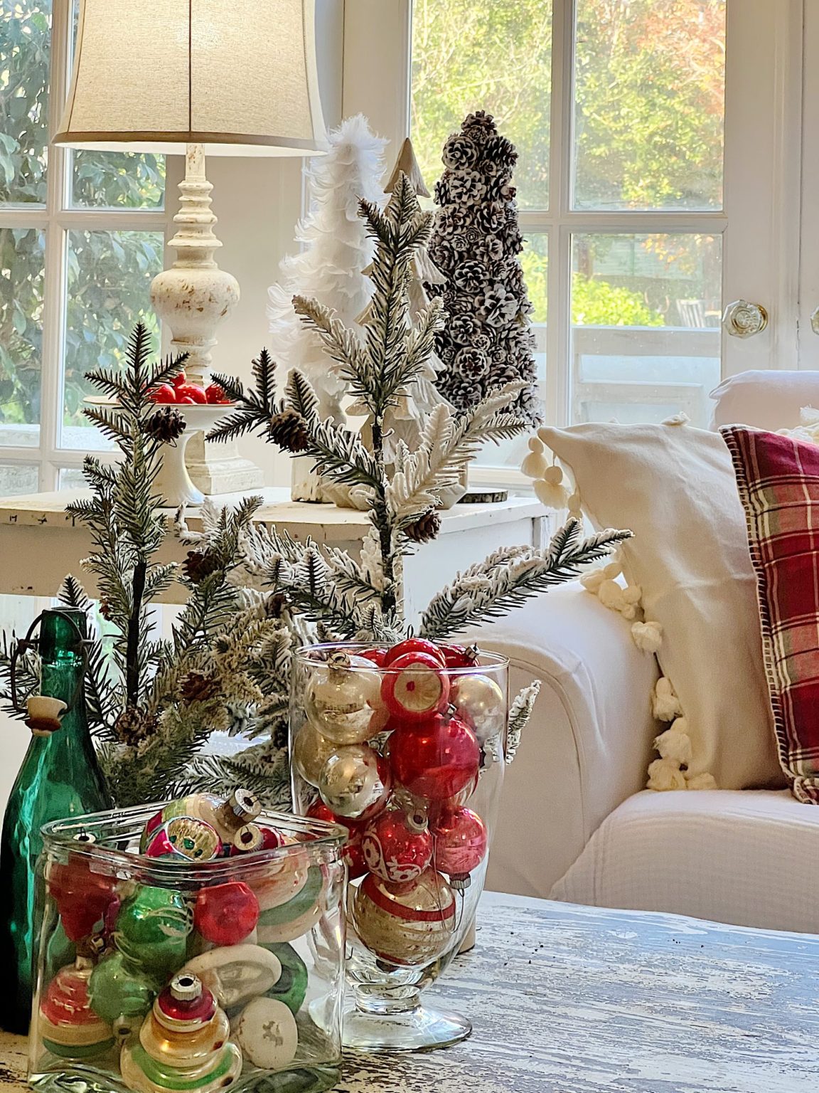 Cozy living room with holiday decorations, including a lamp, small Christmas trees, and glass jars filled with colorful ornaments on a table by a window.