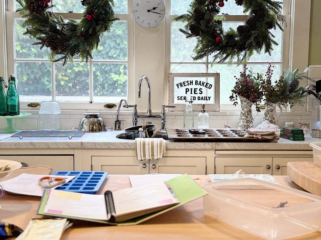 A kitchen with large windows decorated with wreaths. A sign reads "Fresh Baked Pies Served Daily." Books and baking tools are on the counter.