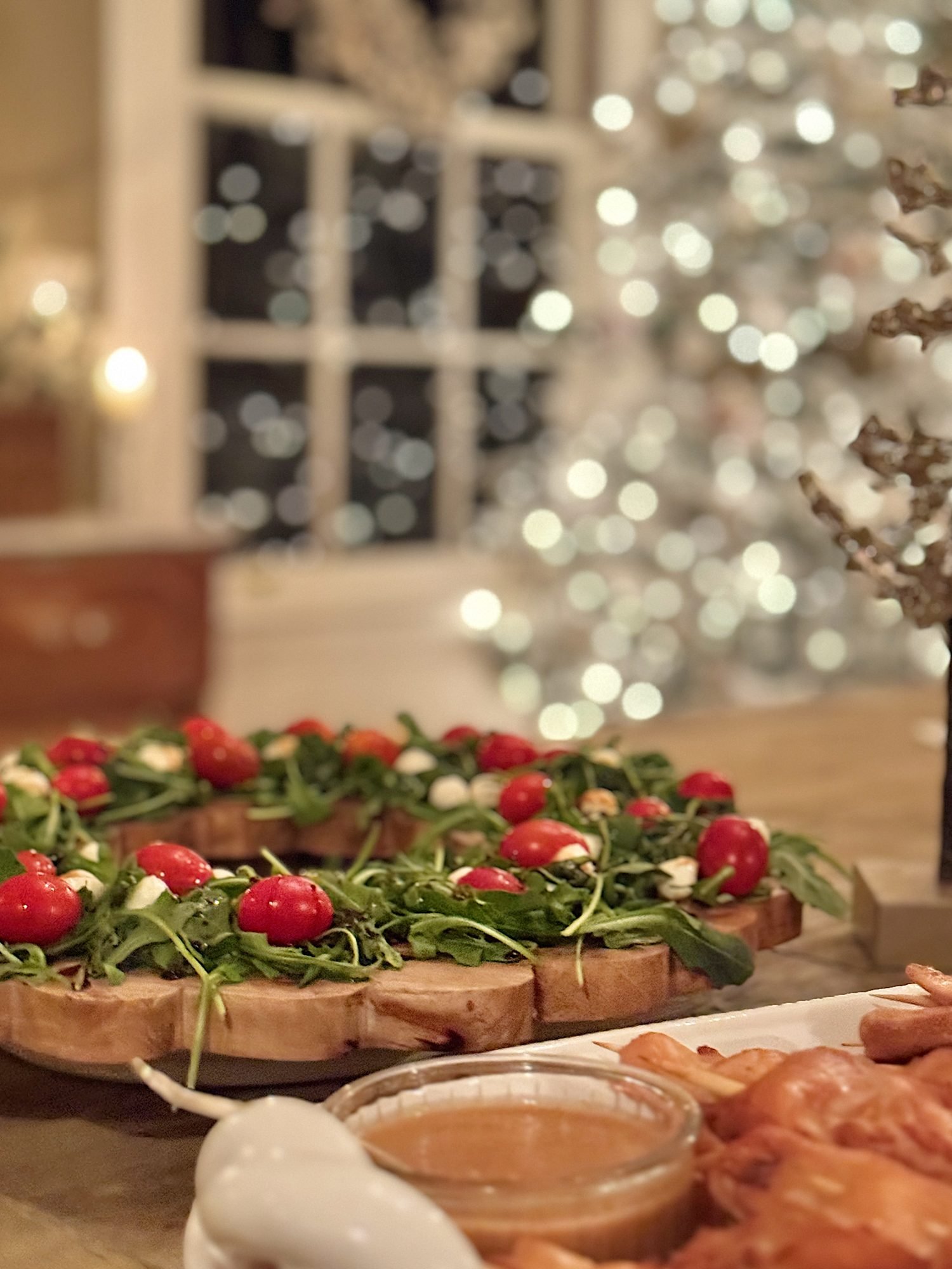 A festive wreath-shaped appetizer made with greens and cherry tomatoes, with a blurred Christmas tree in the background.