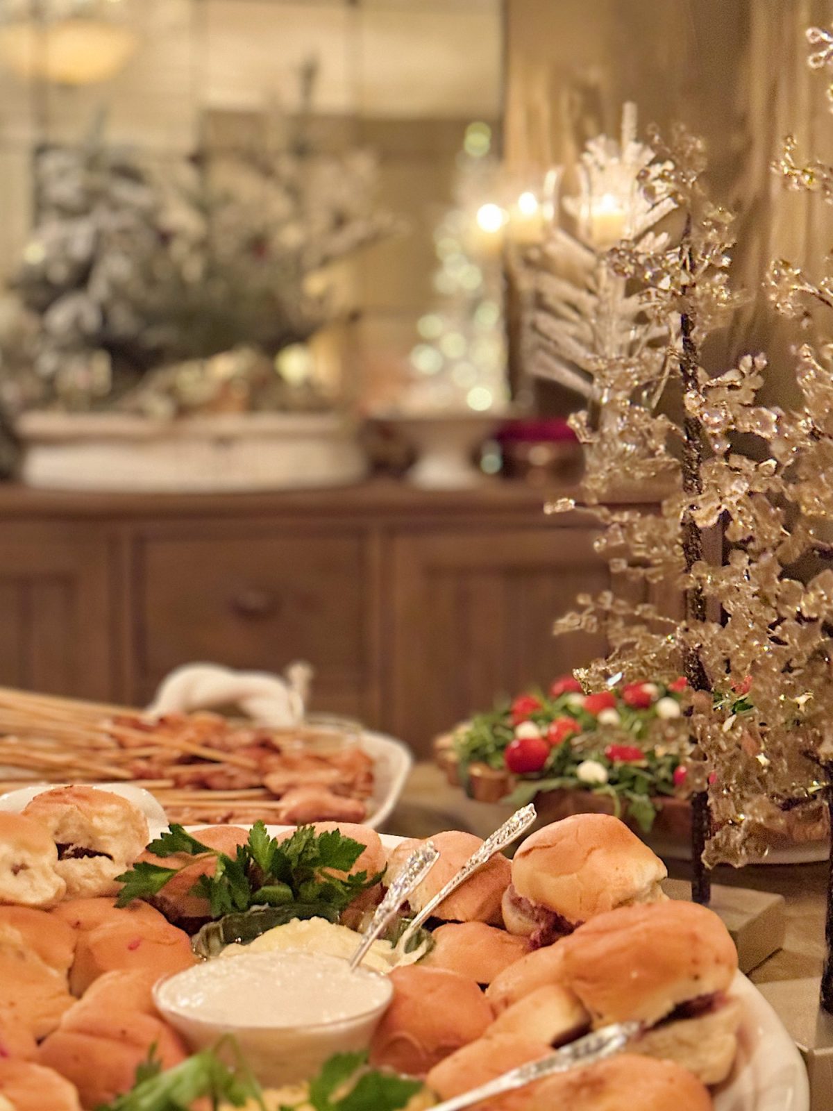 A festive buffet with mini sandwiches, dips, and garnished platters on a wooden table. Blurred background features a decorated, lit Christmas tree.