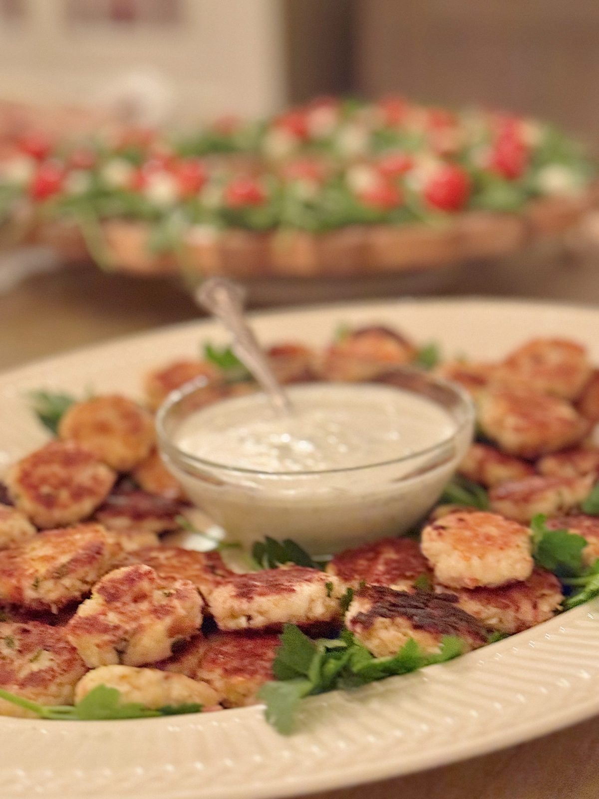 Plate of grilled chicken patties with a bowl of dipping sauce in the center, garnished with herbs. In the background, a platter of appetizers.