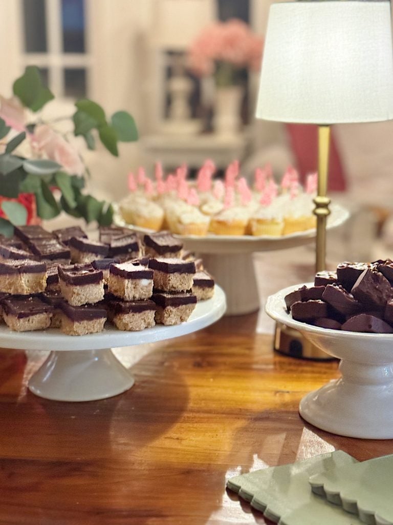 Assorted desserts on cake stands, including chocolate squares and cupcakes with pink decorations, displayed on a wooden table with a lamp.