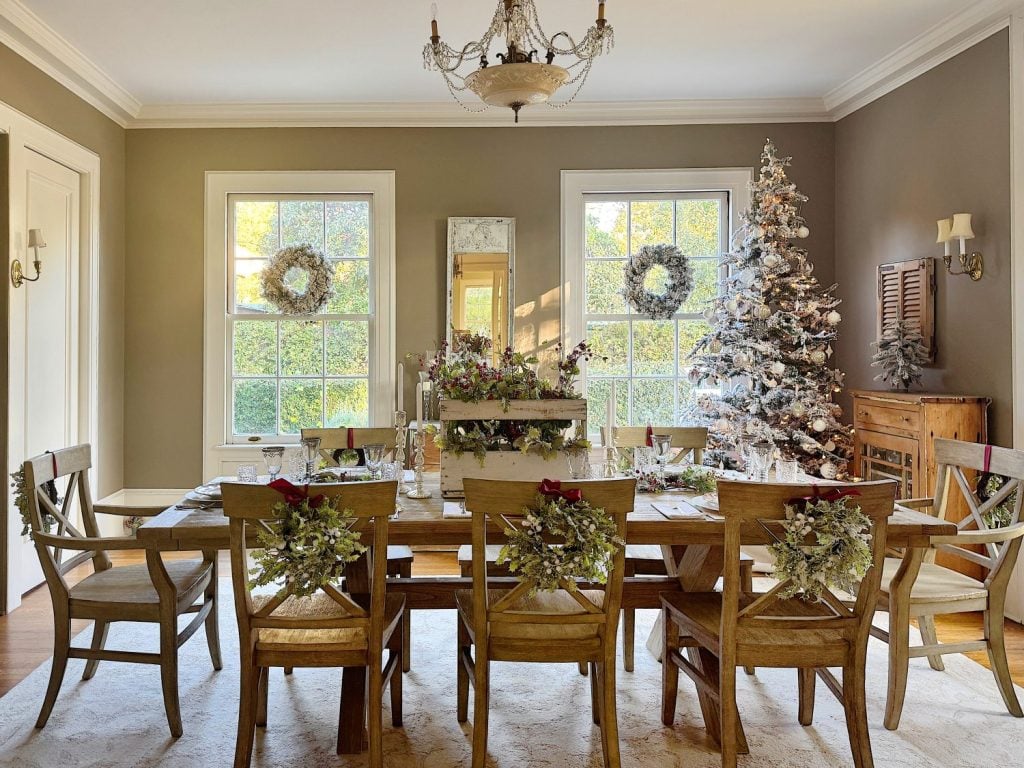 A festive dining room with a decorated table, wreaths on chair backs, and a Christmas tree near the window.