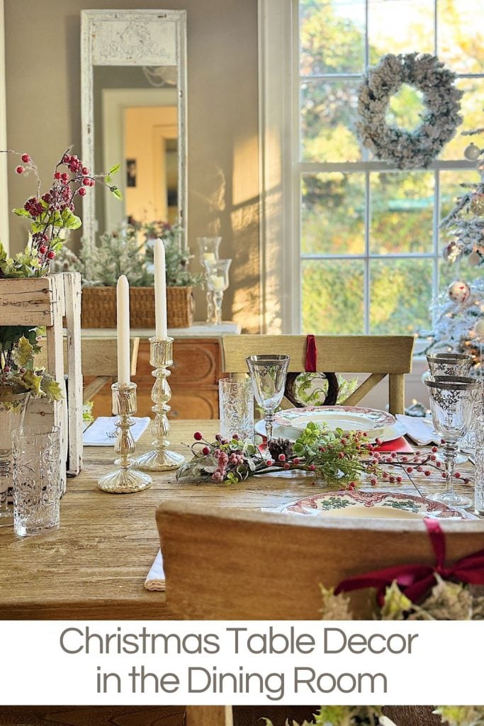 Dining room table with Christmas decor, including candles, glassware, and festive greenery. A mirror and wreath are in the background near a window.