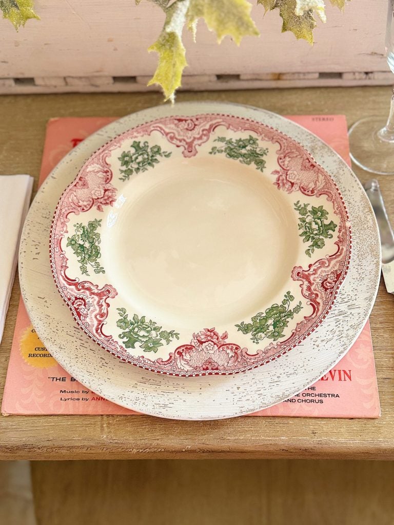 A decorative plate with red and green patterns on a stack of vinyl records, set on a wooden surface with a partially visible glass and napkin.