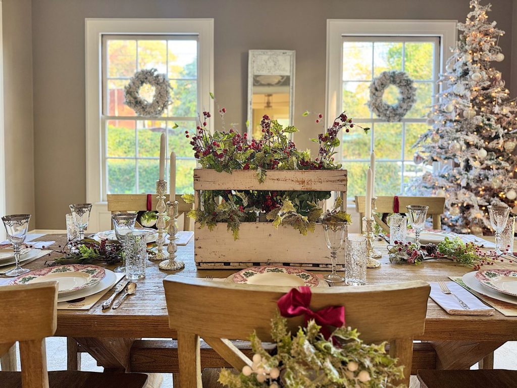 A dining table is set with decorative plates, glasses, and a floral centerpiece. A decorated Christmas tree and wreaths are visible in the background near large windows.
