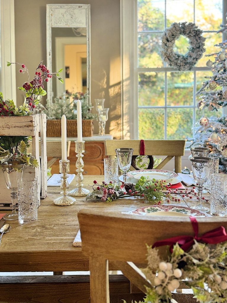 A festive dining table set with crystal candlesticks, floral arrangements, glassware, and wreath decorations in a room with a decorated Christmas tree and large window.