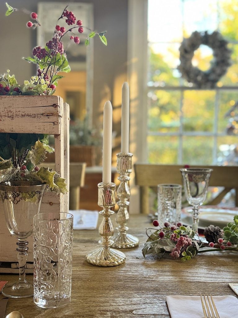 Dining table with ornate glassware, two candlesticks, floral decorations, and a rustic wood box centerpiece. Sunlight filters through the window in the background.