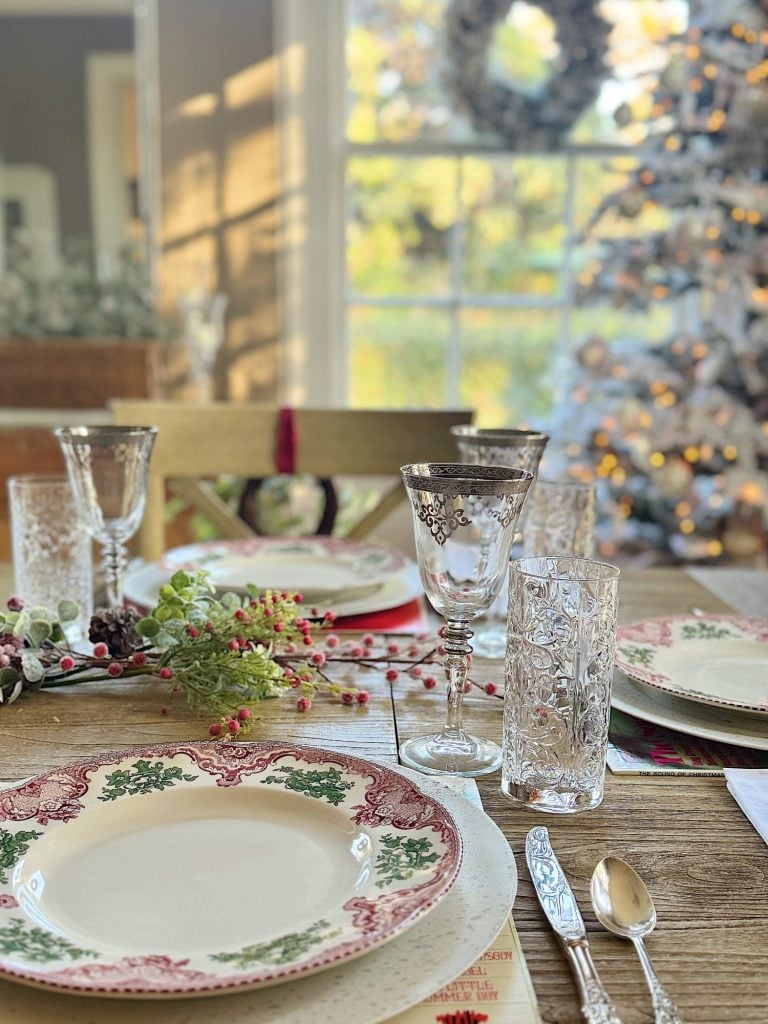 A dining table set with Christmas-themed plates, glassware, and cutlery. A decorated Christmas tree is visible in the background near a window.