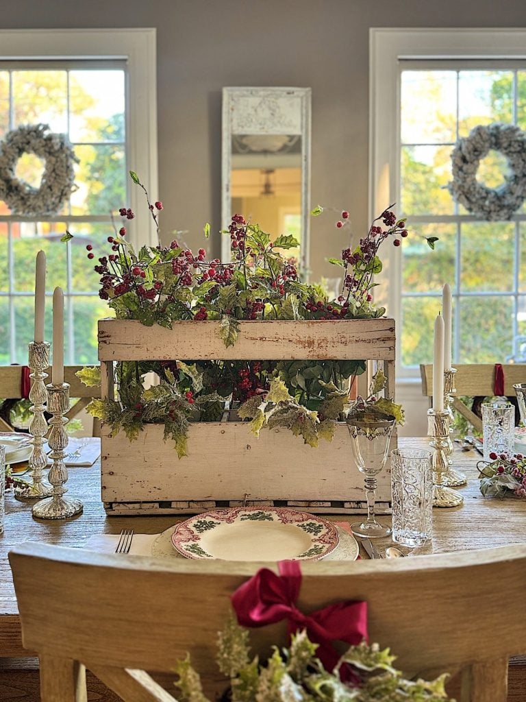 Dining table with festive decor, featuring a wooden crate with red berries and greenery, crystal candle holders, and decorative plates. Two wreaths hang on the windows in the background.