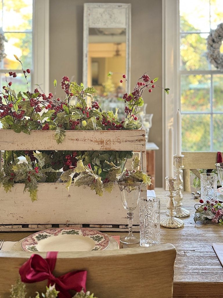 Festive dining table with floral centerpiece, glasses, candles, and Christmas-themed plates, set in a room with large windows and wreaths on the walls.