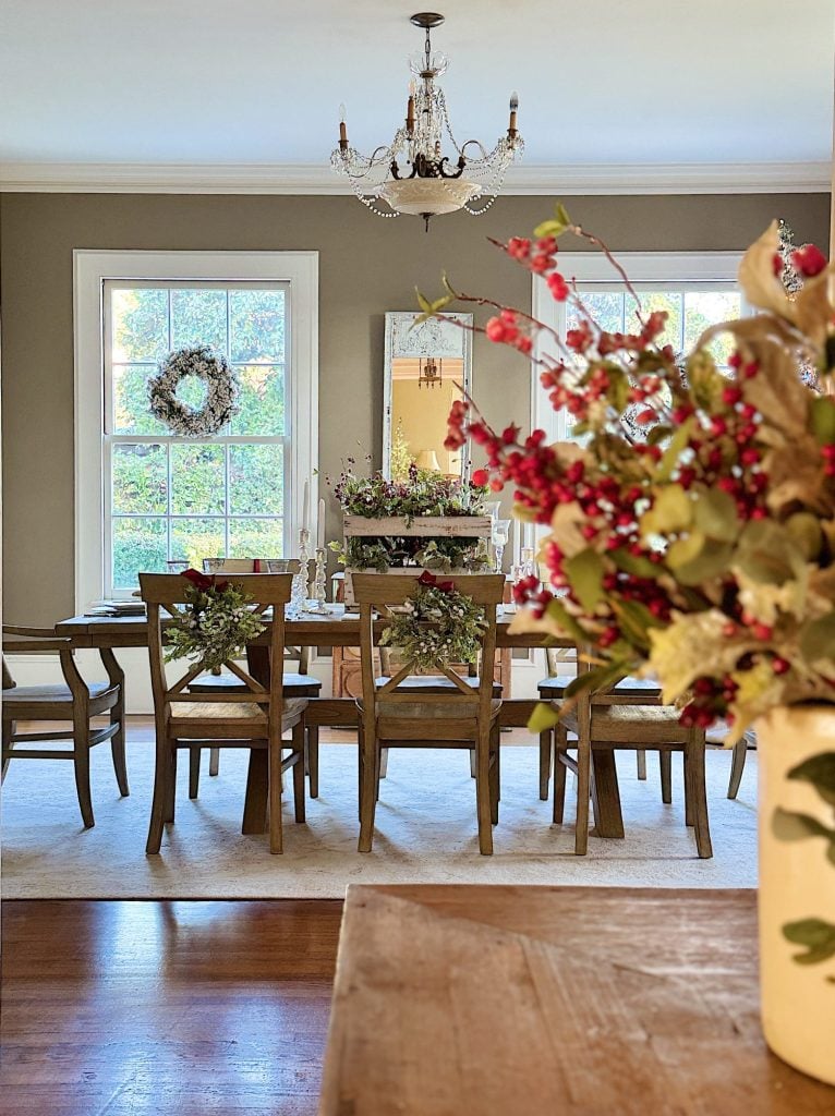 A dining room with a wooden table and chairs, decorated with wreaths. A chandelier hangs above, and a floral arrangement is in the foreground. Large windows show greenery outside.