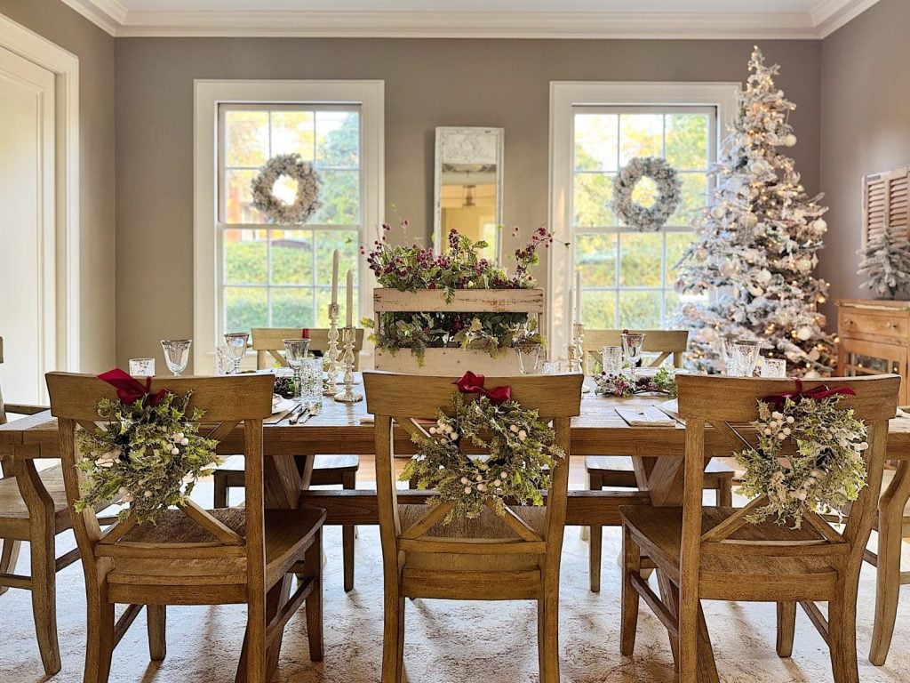 Wooden dining table set with festive decorations and floral arrangements. Chairs have wreaths on the back. Christmas tree and wreaths by the window.