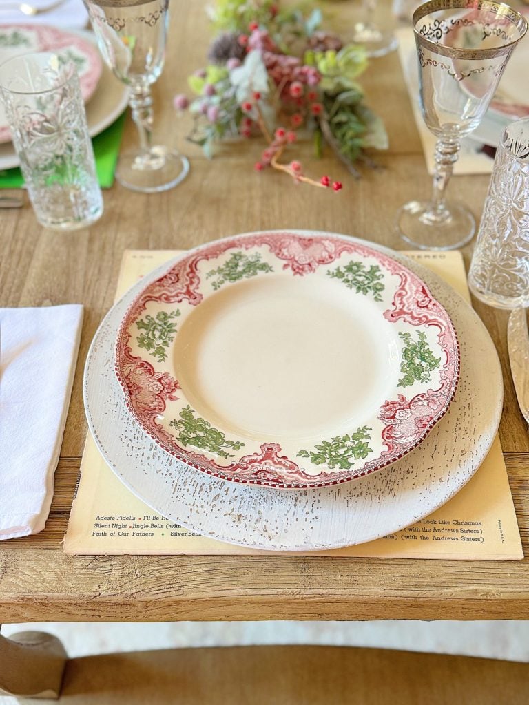 A dining table set with vintage-style plates, crystal glasses, and a floral centerpiece on a wooden surface.