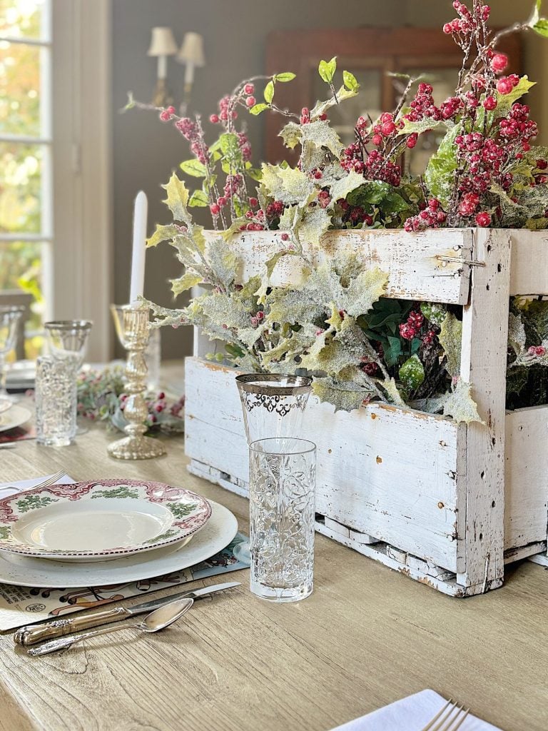 Table setting with a white wooden box centerpiece containing frosted greenery and red berries. Elegant glassware and patterned plates are placed on the wooden table.