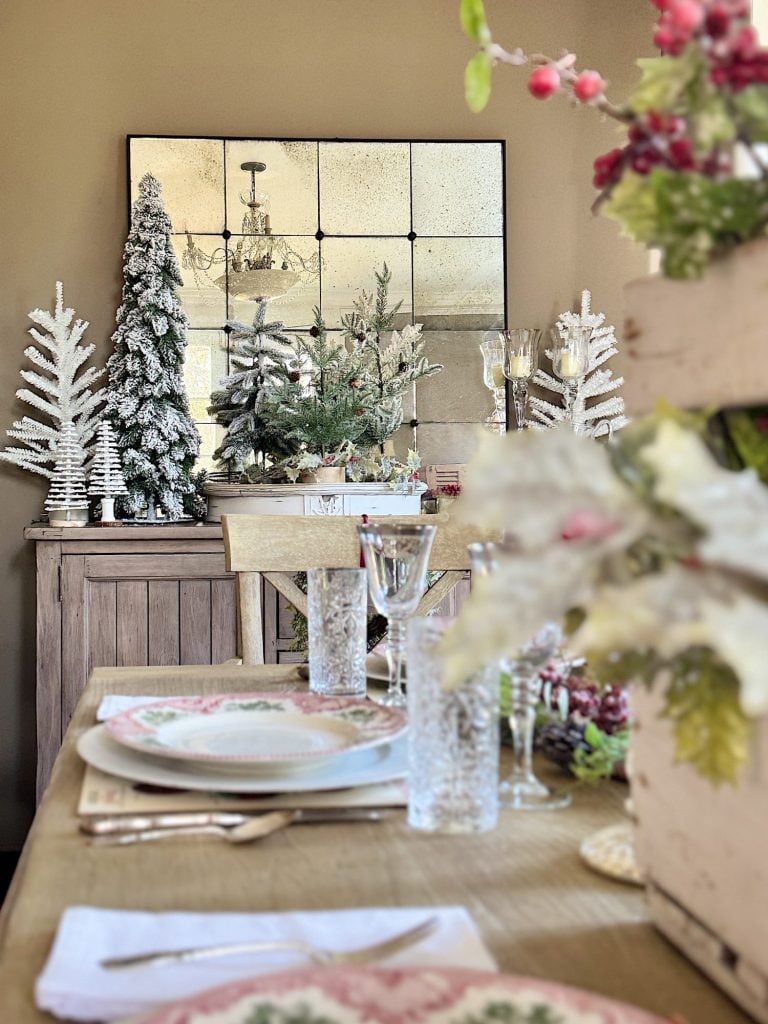 Festive dining table with elegant dinnerware, glassware, and holiday decorations, including small Christmas trees and candles. A large mirror with a rustic frame hangs on the wall in the background.