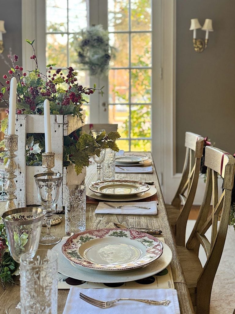 Elegant dining table set for four with floral plates, crystal glasses, white candles, and a decorative centerpiece, situated near large glass doors with a view of greenery outside.