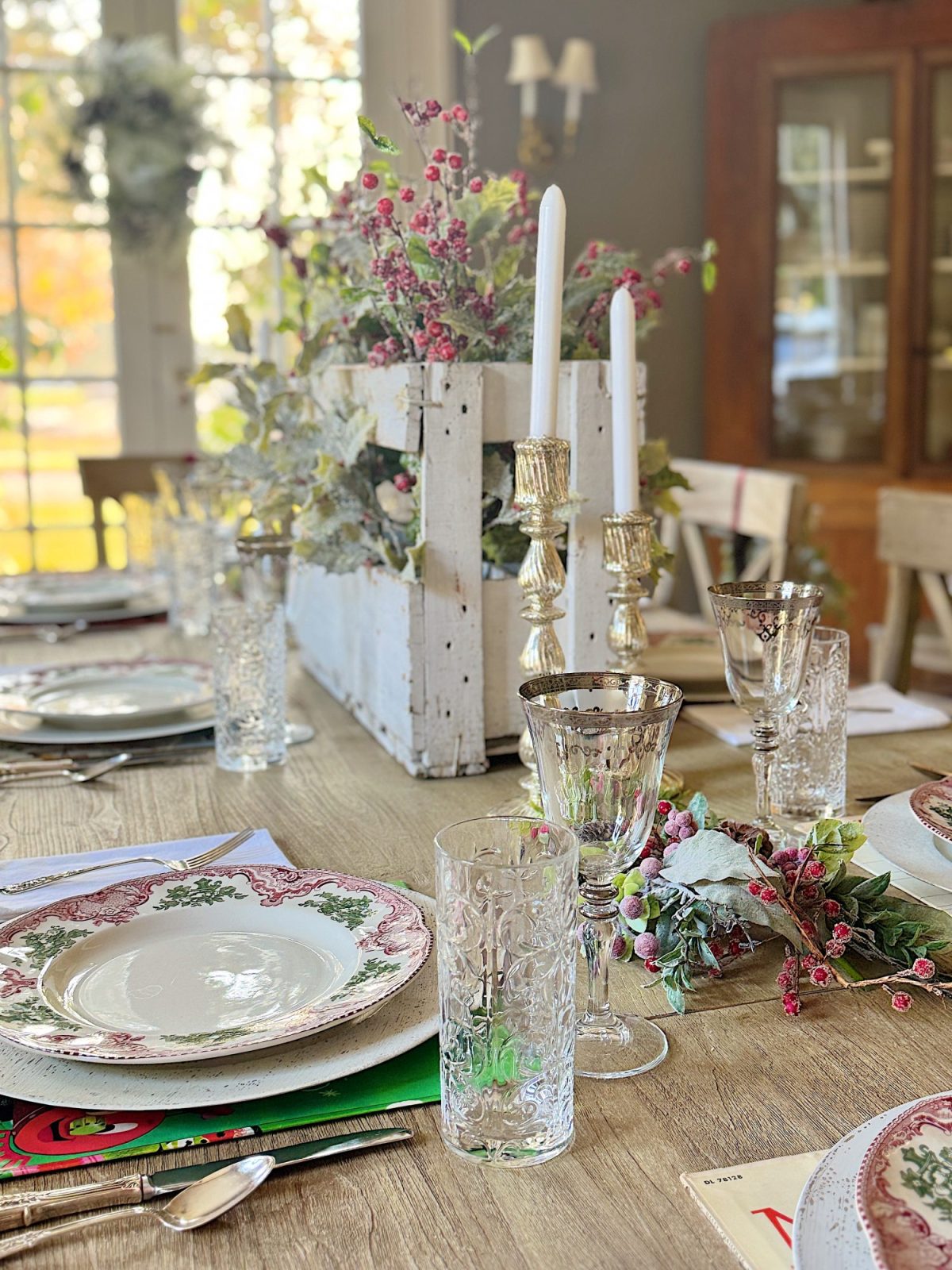 A dining table set for a meal with floral-patterned plates, clear glassware, and a centerpiece featuring candles and a rustic white box with faux flowers and foliage.