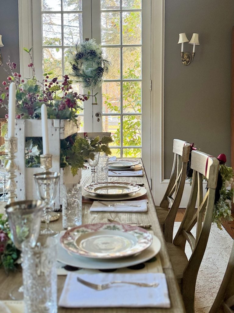 A dining table set for a meal, featuring plates, cutlery, glasses, and candles. The table is decorated with floral arrangements. French doors with a wreath are in the background.