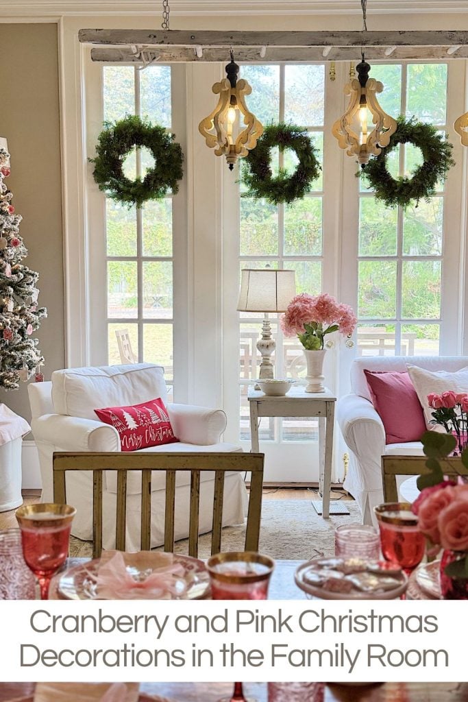 Family room with Christmas decorations. Two white chairs, pink and red cushions, festive wreaths, and a Christmas tree. Table with pink flowers and gold-rimmed glasses.