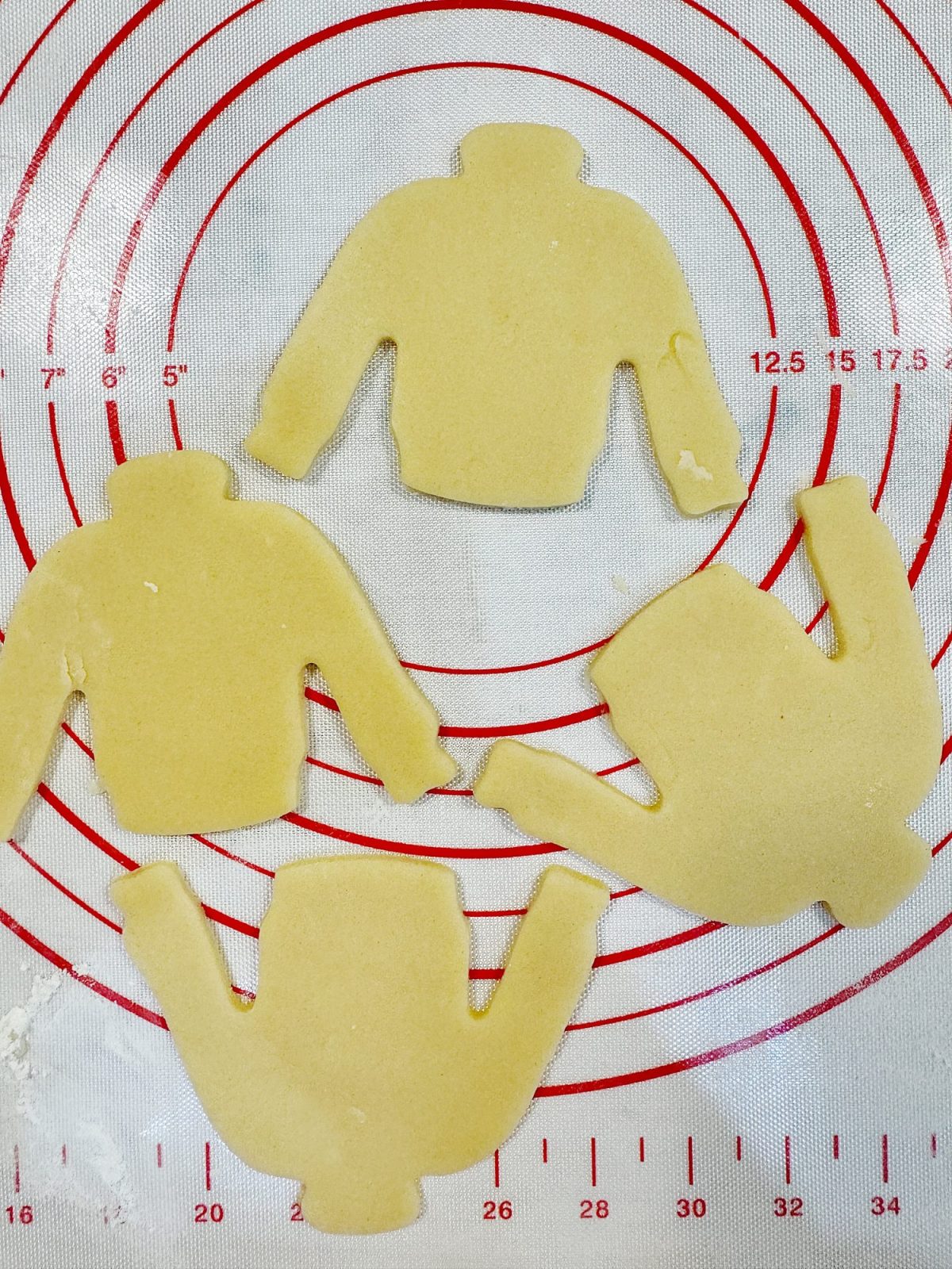 Four sweater-shaped cookie dough cutouts on a red and white circular measurement mat.