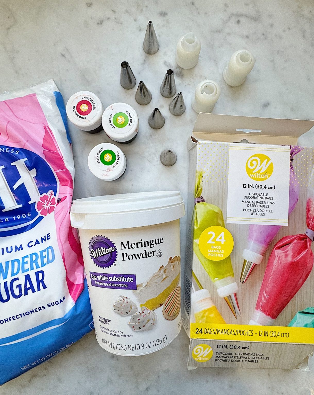 Baking supplies on a marble surface, including powdered sugar, meringue powder, food coloring jars, piping tips, and a box of decorating bags.