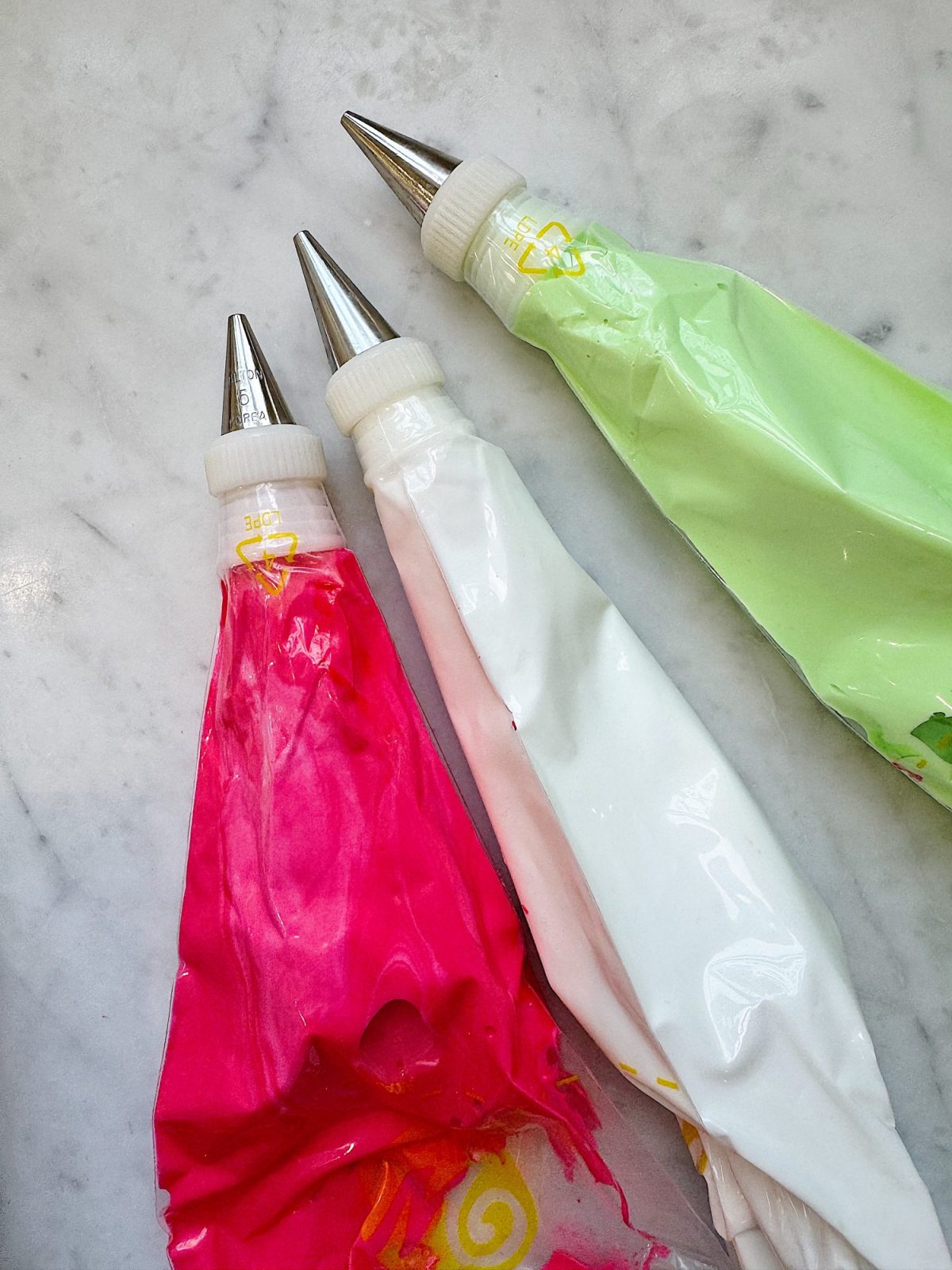 Three piping bags with metal tips, filled with pink, white, and green icing, lie on a marble surface.