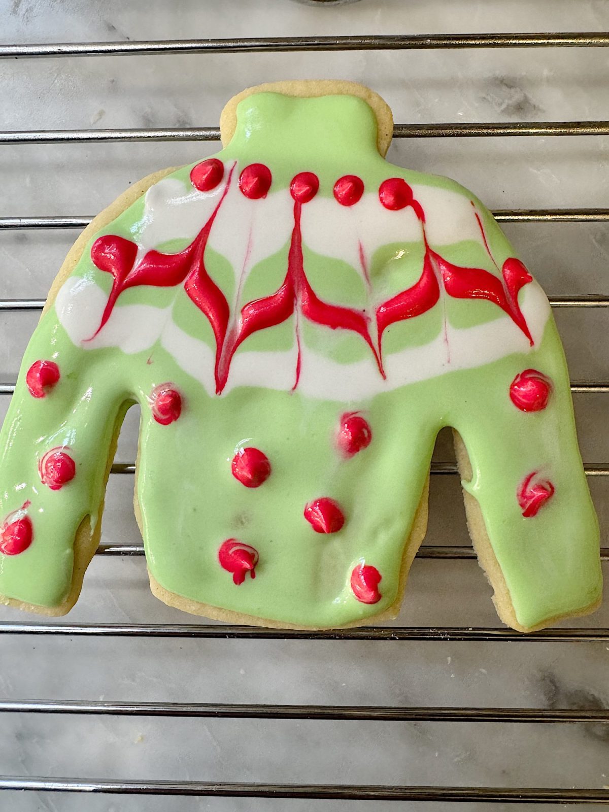 A sweater-shaped cookie with green icing, red and white patterns, and red dot decorations on a wire rack.