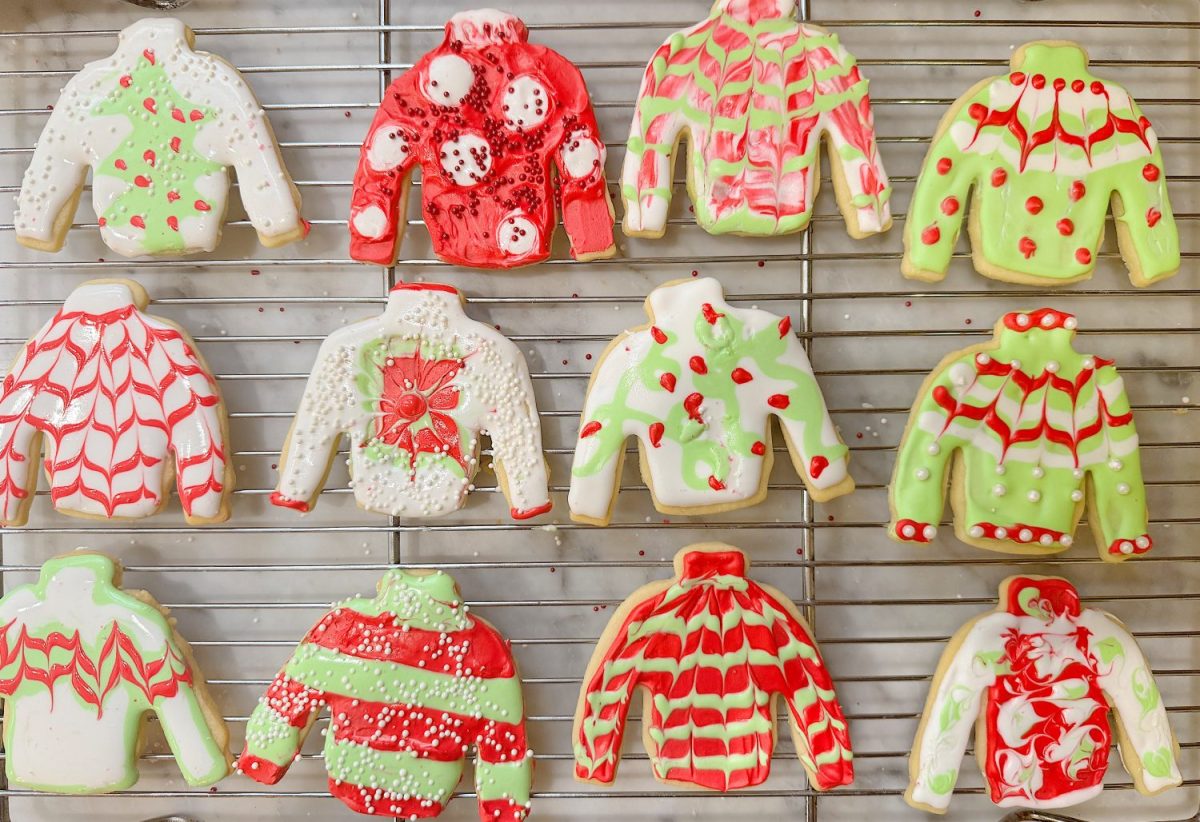 Twelve sweater-shaped cookies with various red, green, and white icing designs are arranged on a cooling rack.