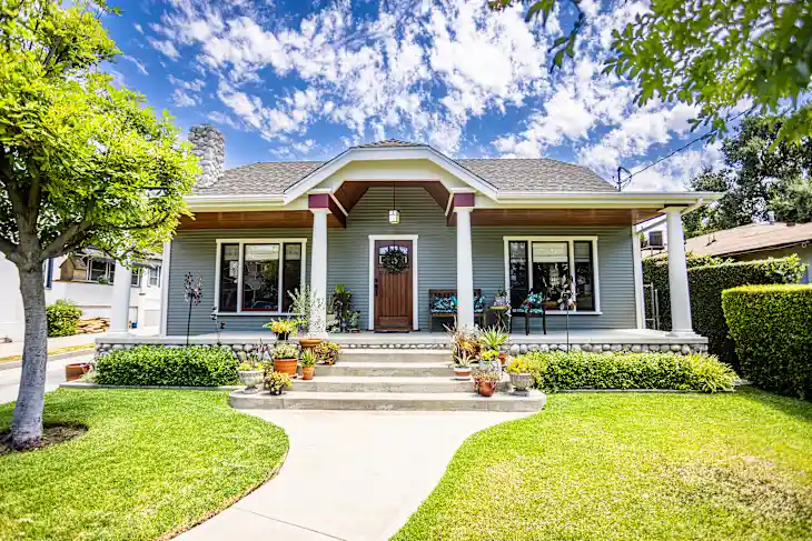 A gray bungalow-style house with a wooden front door, manicured lawn, potted plants, and a concrete pathway under a partly cloudy sky.