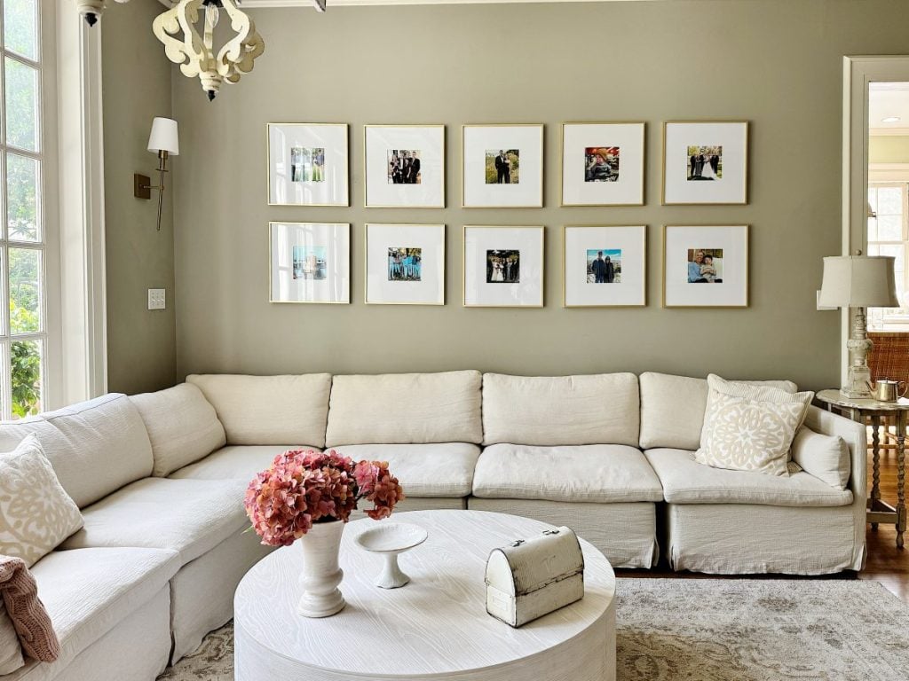 A living room with a white sectional sofa, round coffee table with flowers, and nine framed photos arranged in a grid on a light olive wall showcases stylish home decor items.