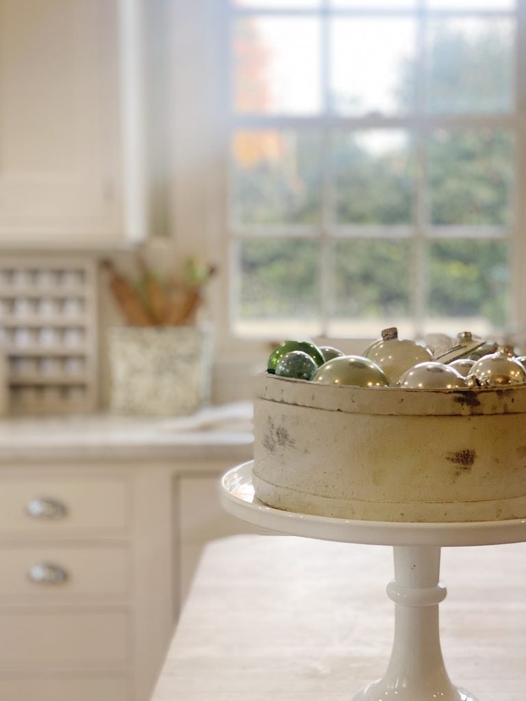 A cake stand holds a vintage tin filled with assorted Christmas ornaments. In the background, a kitchen counter and window with a blurred view of greenery are visible.