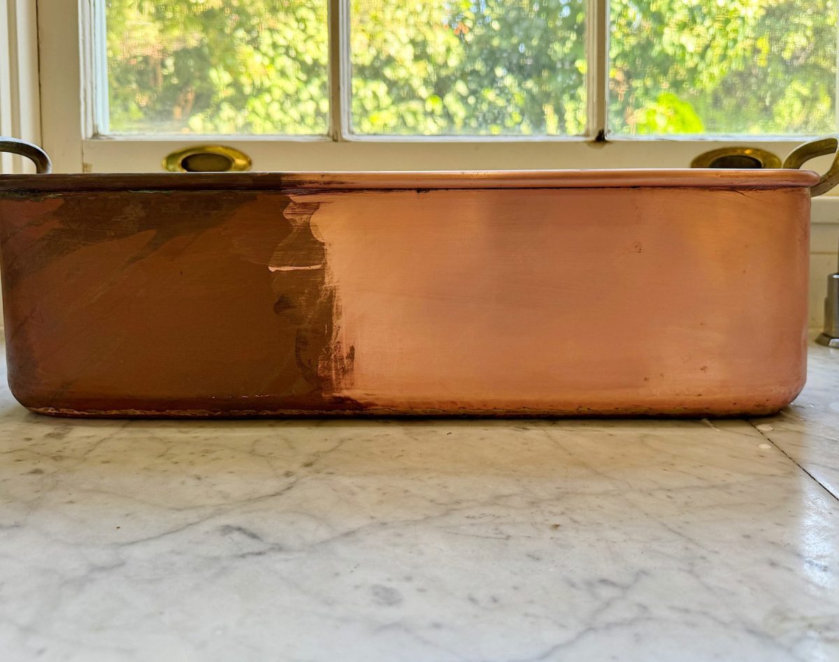 A copper pan sits on a marble countertop, partially polished to reveal a bright, shiny surface beside an unpolished, tarnished section—perfect inspiration for unique table setting ideas. A window and greenery are in the background.