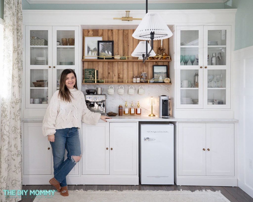 A woman in a white sweater and jeans stands by a home coffee bar with a small fridge, coffee machine, mugs, and decor on white cabinetry and wooden shelves.