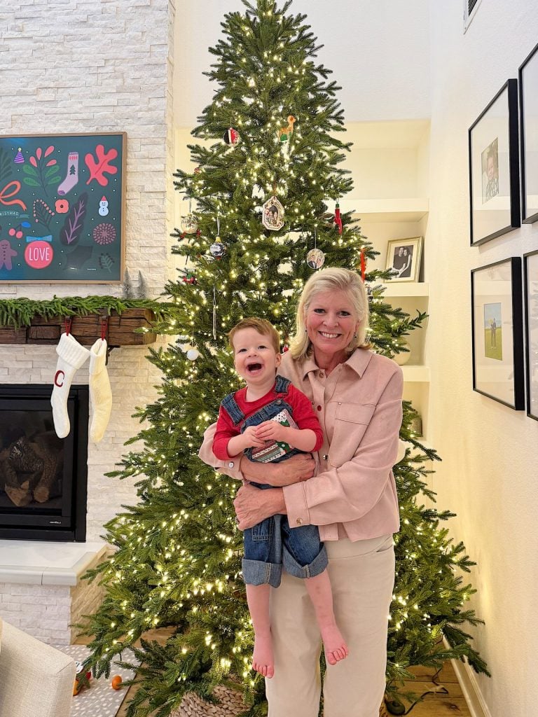 A woman holding a smiling toddler in front of a decorated Christmas tree with lights. There is a stocking hanging by the fireplace and artwork on the wall.