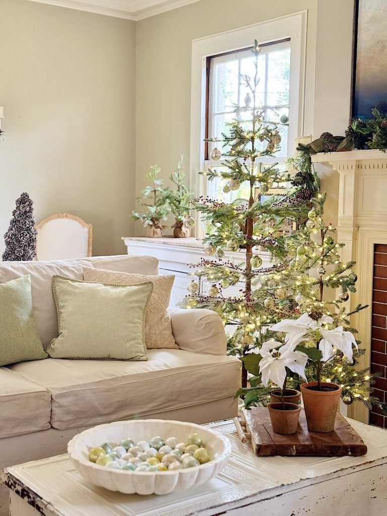 Cozy living room with a lit Christmas tree, white couch with green pillows, a fireplace, and decorative items including potted plants and a bowl of pastel ornaments.