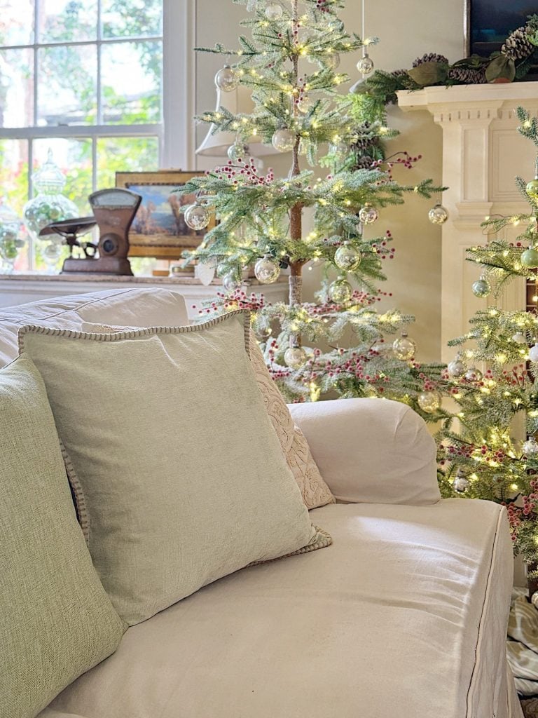 A cozy living room decorated for Christmas with lit trees, ornaments, and a beige couch with green and beige pillows. Light filters through a window in the background.