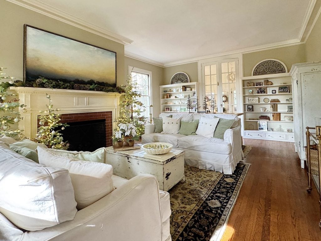 A living room with white sofas, a fireplace, decorative shelves, and a large painting above the mantel. The room features wooden floors and light-colored walls.