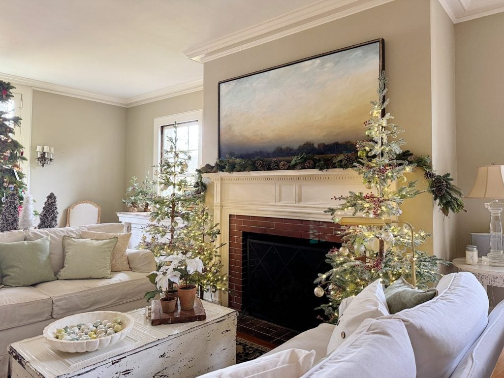 A cozy living room decorated for Christmas with two lit trees by the fireplace, soft couches, and a large landscape painting above the mantel.