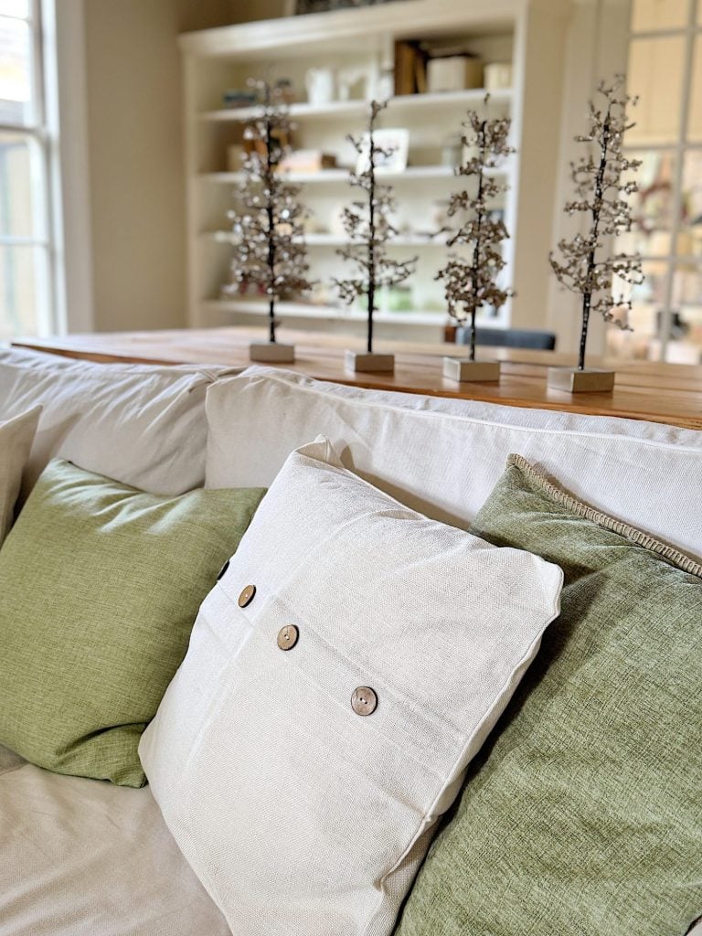 A cozy sofa with white and green pillows is positioned in front of a wooden shelf displaying decorative metal tree sculptures.