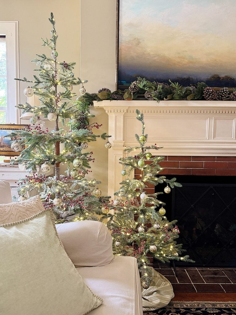 Three decorated Christmas trees with lights and ornaments stand near a white fireplace adorned with greenery and pinecones. A white couch is in the foreground.