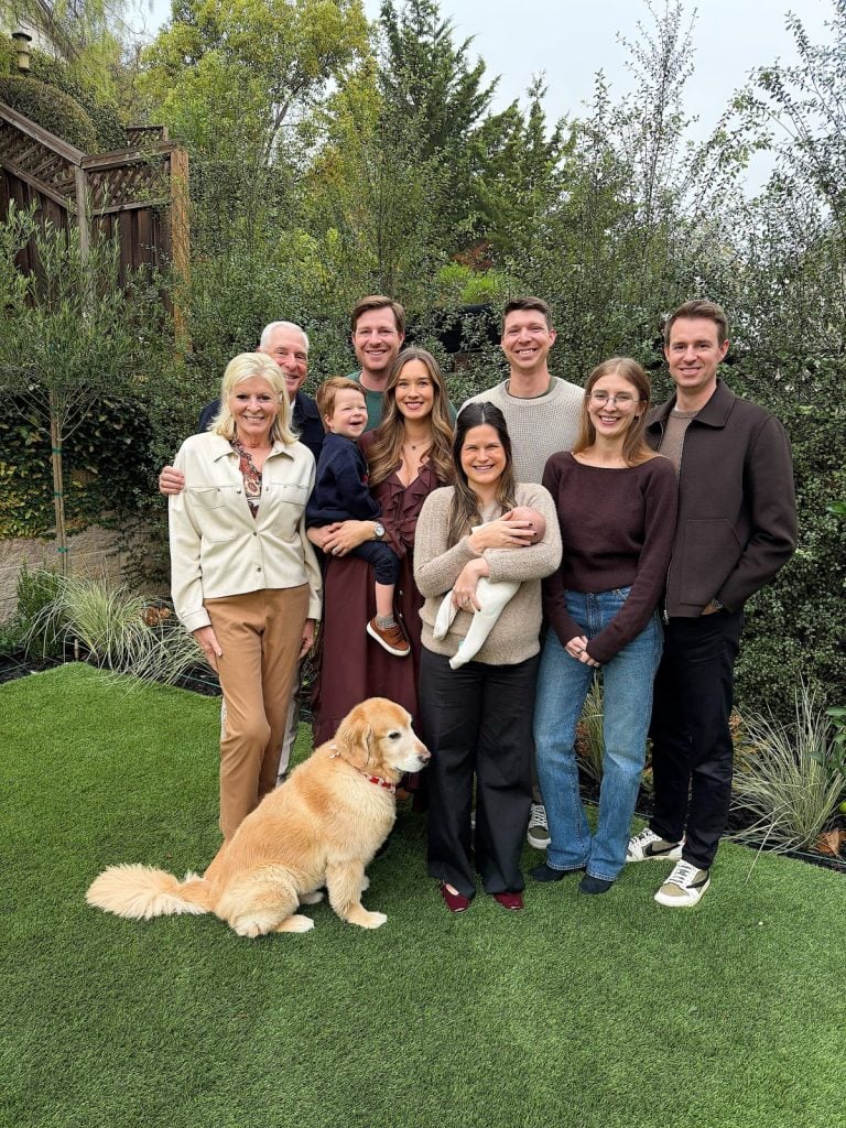 A group of nine people, spanning multiple generations, and a golden retriever pose together outdoors on green grass, surrounded by trees and bushes, sharing a cheerful moment perfect for a Merry Christmas greeting.