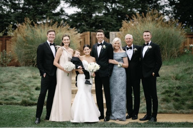 A wedding party poses outdoors on grass, with six adults in formal attire and one child. The group stands in front of wooden fencing and tall grass.